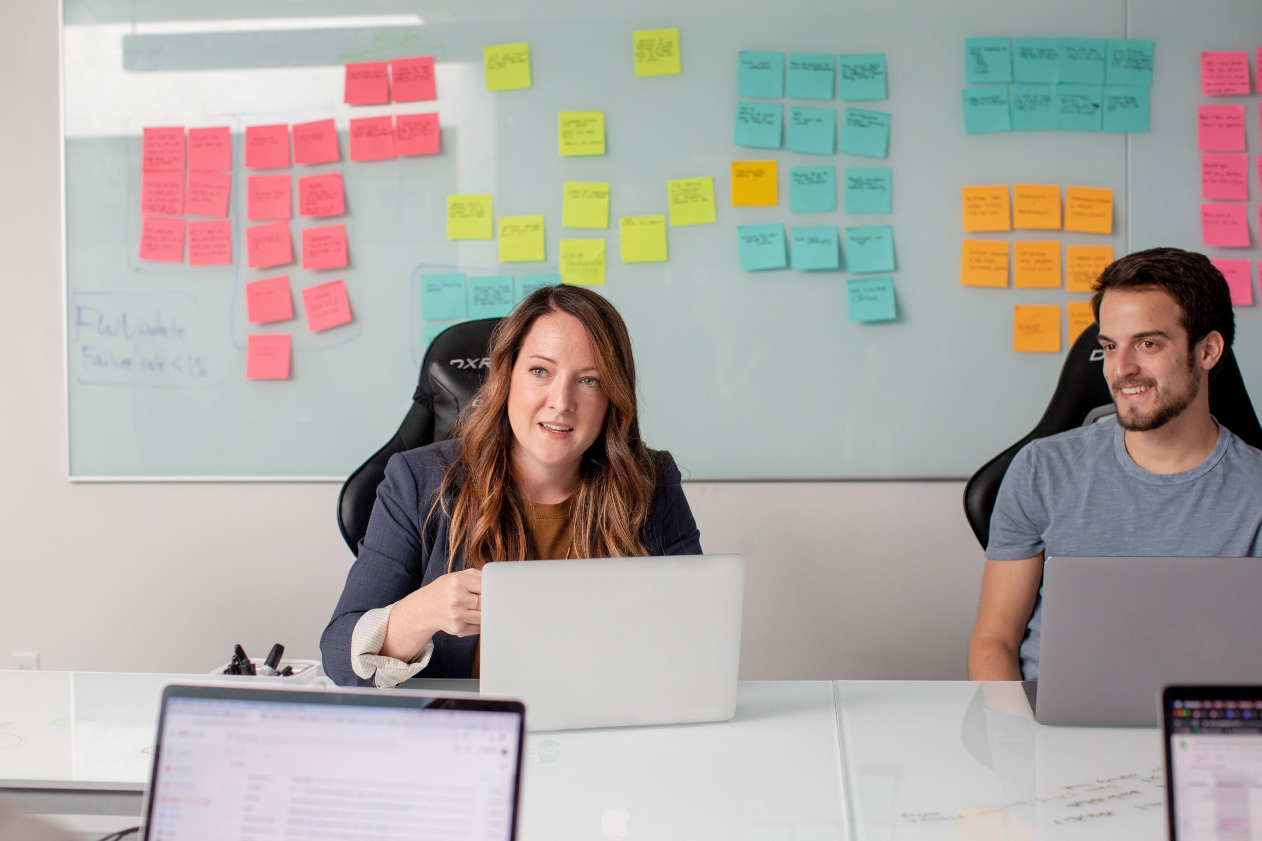 A man and woman sitting at their laptops. Various colored sticky notes line the whiteboard behind them.