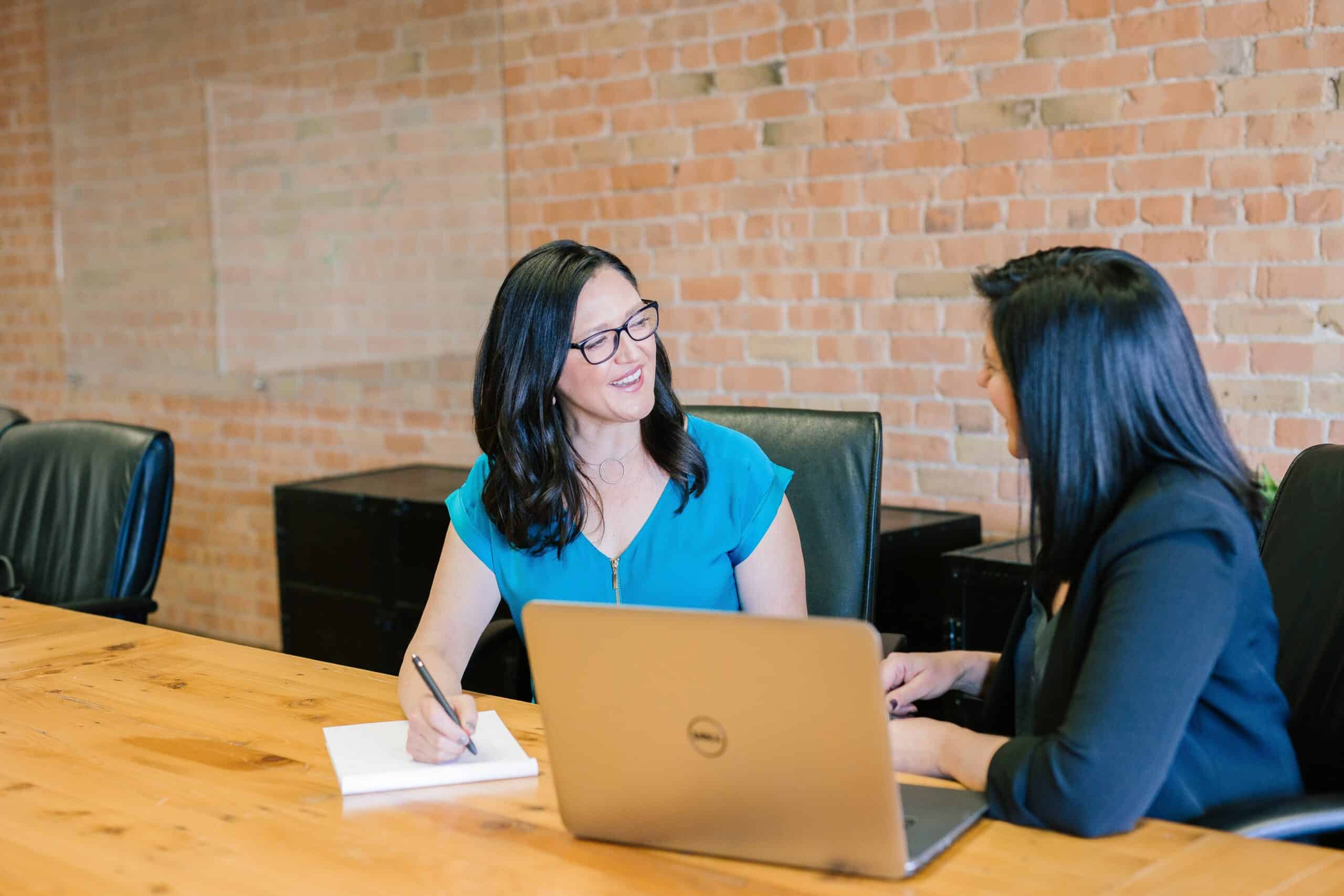 Woman in a teal shirt sitting in front of an Apple laptop. Talking to a lady in black.