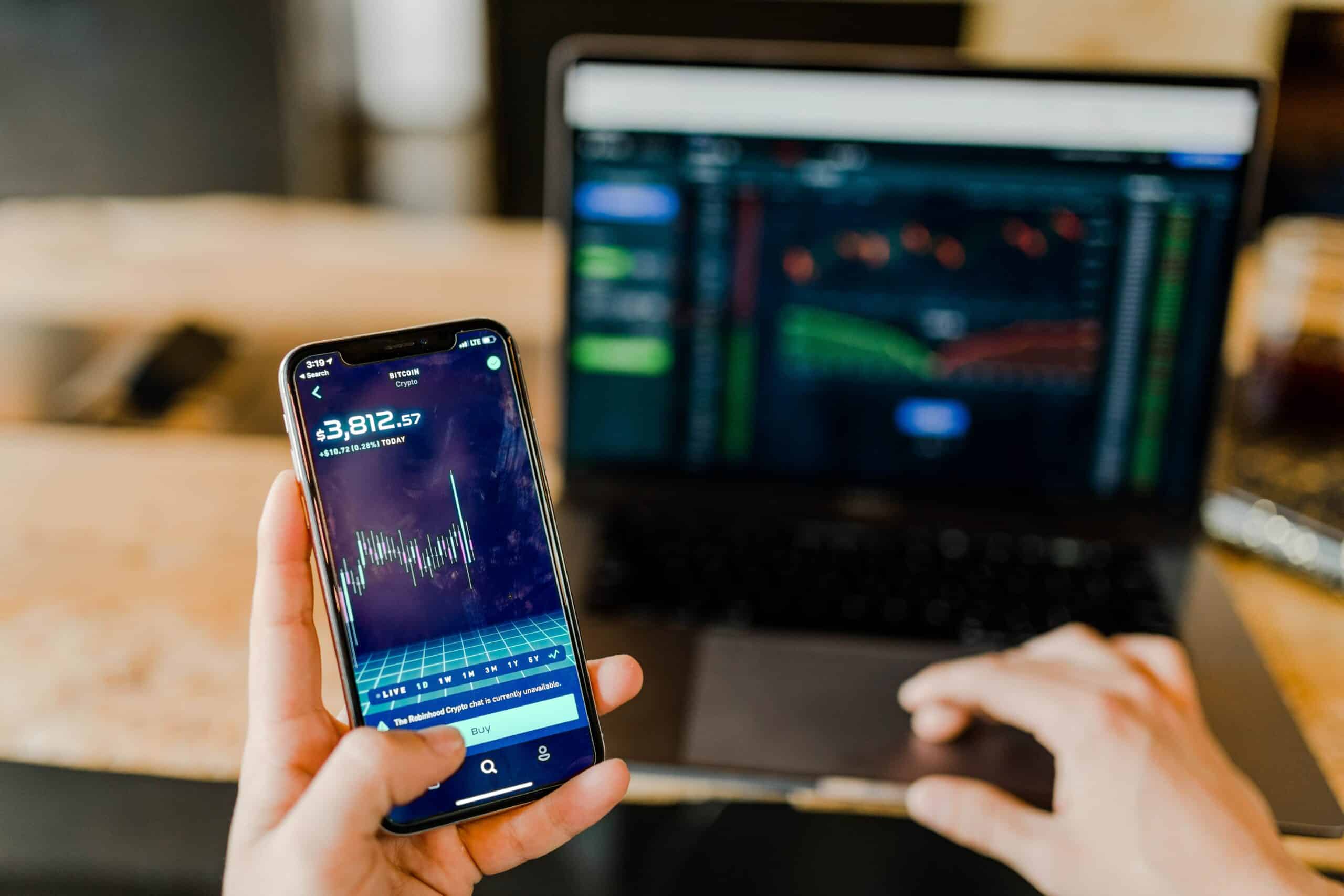 Person holding a cell phone while sitting in front of a laptop. Looking at financial market information