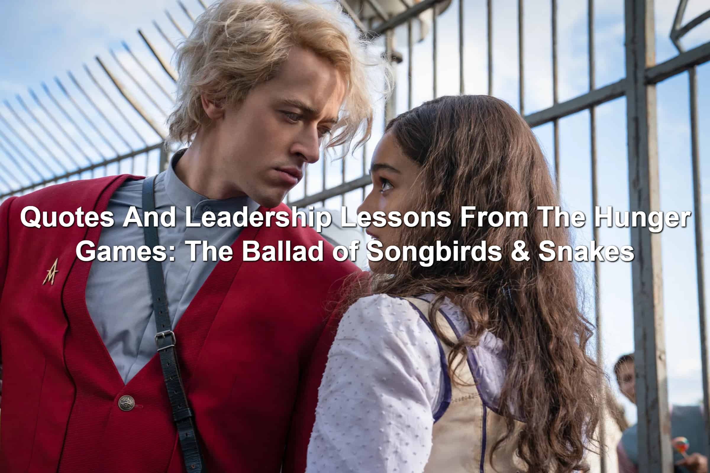 Scene from The Hunger Games: The Ballad of Songbirds and Snakes. Tom Blyth and Rachel Zegler. Blonde man in a red suit coat looking intently at a girl with brown hair.